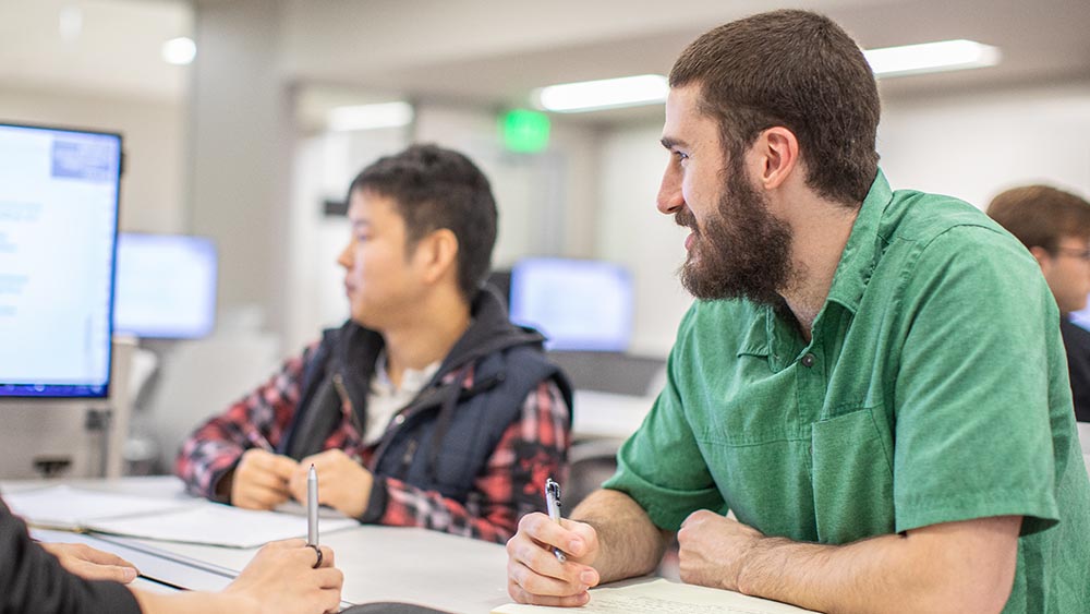 一名穿着绿色衬衫的男学生拿着一支笔看着监视器。另一名学生出现在背景中。