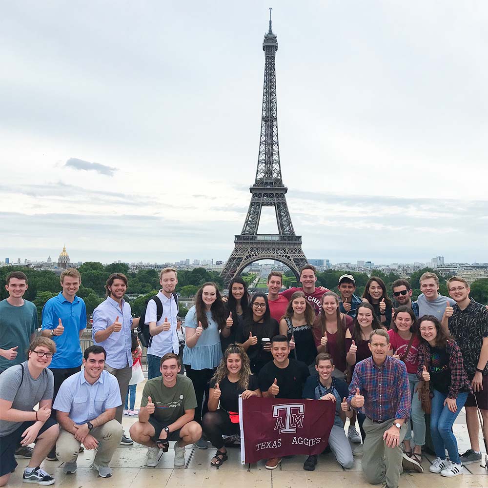 一大群海外留学的学生和一名竖起大拇指的教师，以及在埃菲尔铁塔前摆姿势的德州Aggies旗。