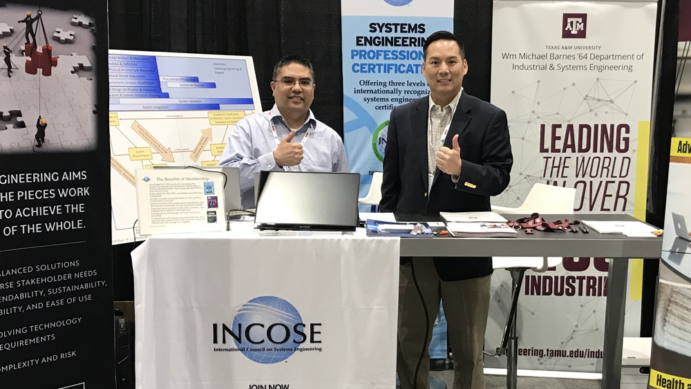 两名男子站在INCOSE会议的桌子后面。他们周围贴着工业与系统工程标志和INCOSE标志的海报。
