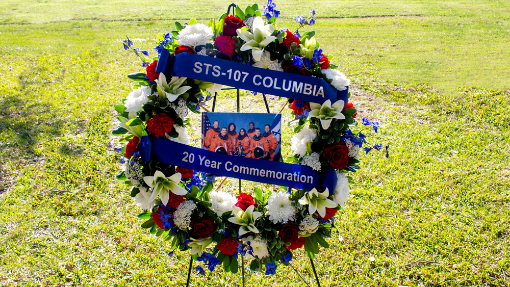 公园里立着一个花圈。花圈中间是七名宇航员的照片。蓝色丝带上写着“STS-107哥伦比亚20周年纪念”。