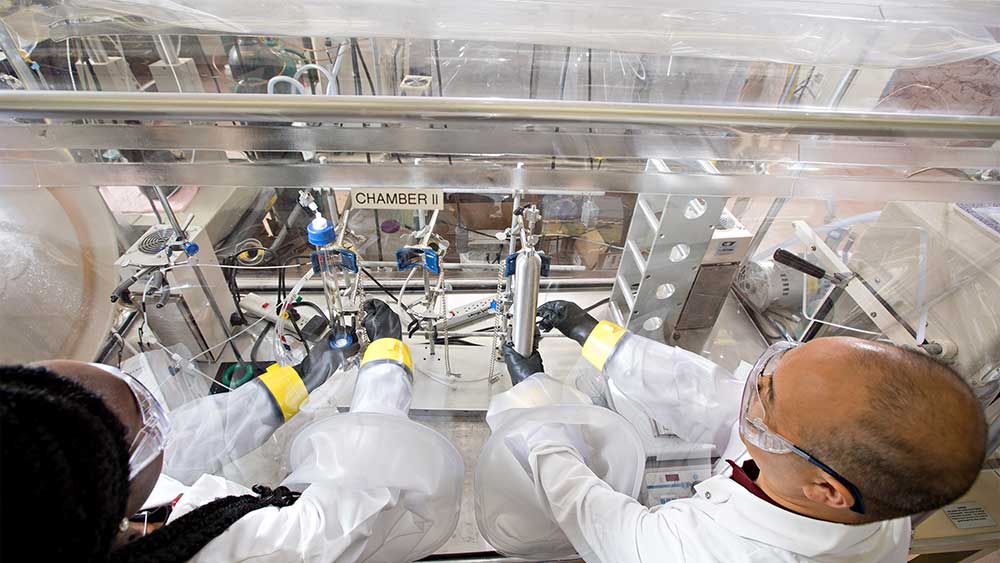 男女研究人员穿着实验室大褂和其他安全装备，在封闭的实验站工作